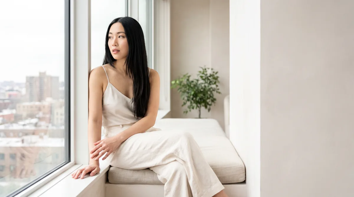 A woman sitting at a window seat looking away mid-thought, nothing in her hands
