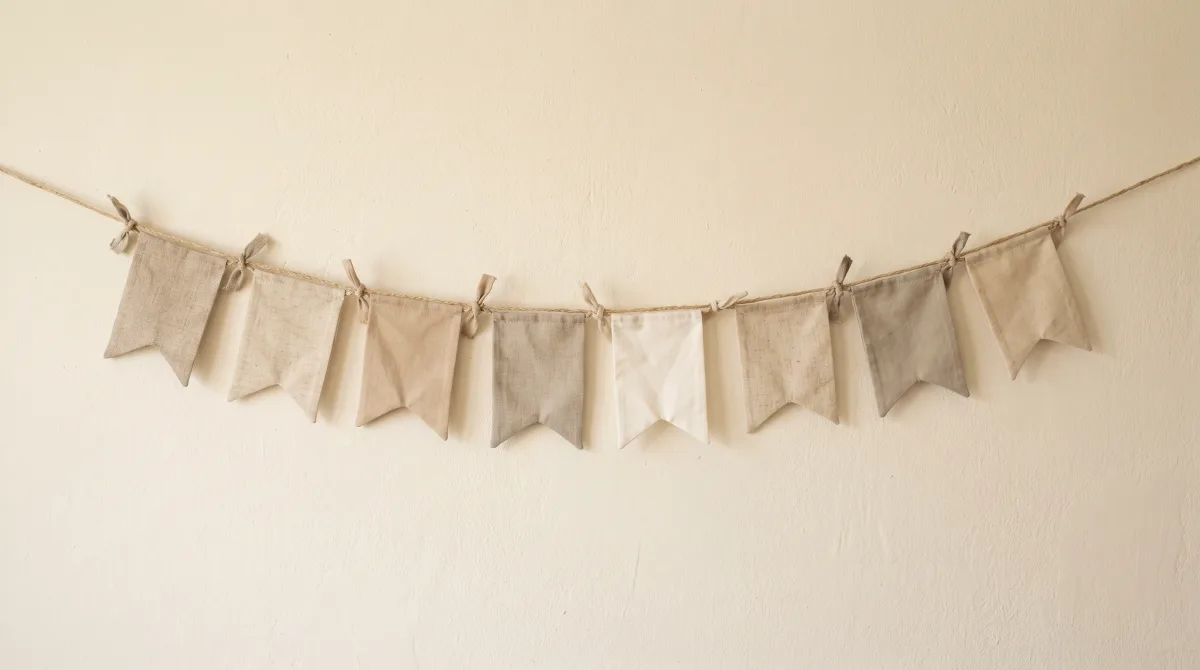 A row of small fabric pennants in nearly identical shades of beige and oatmeal, pegged on a line against warm plaster, one flag very slightly off-white