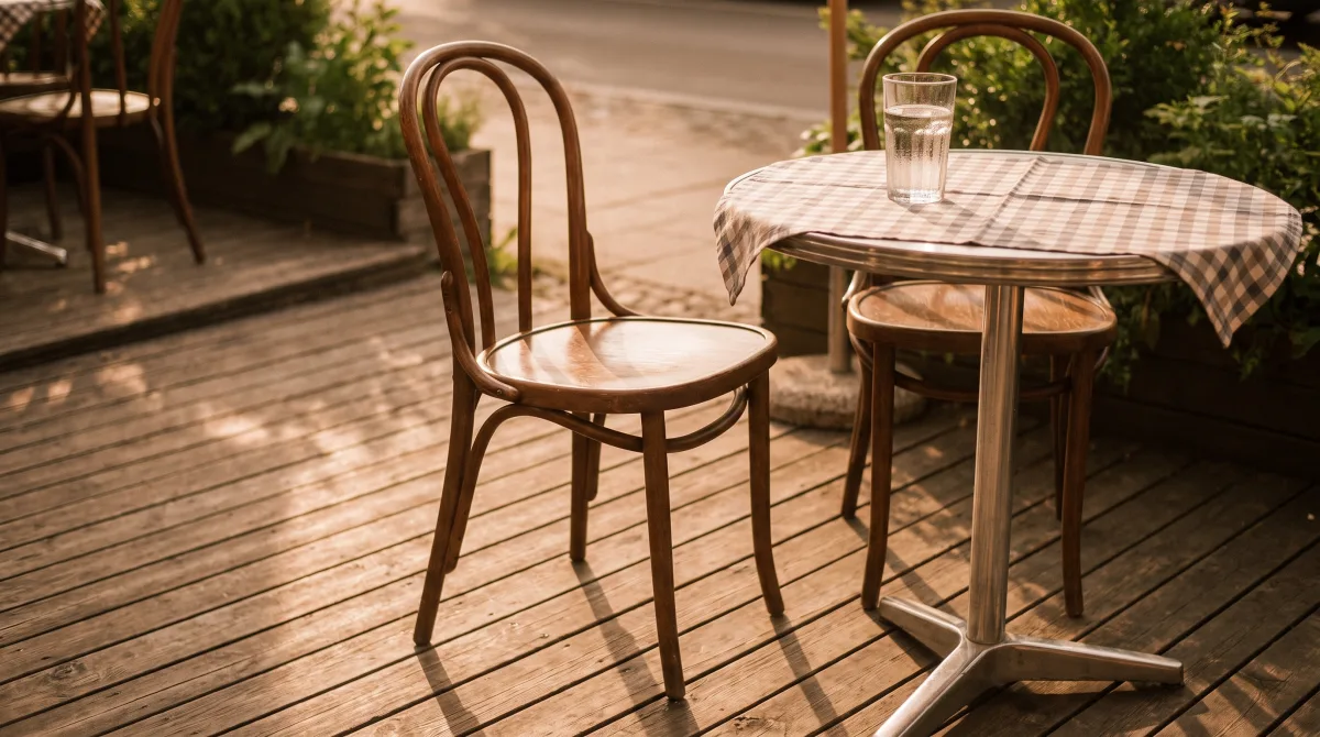 A single café chair pulled out at an empty two-top table, one untouched glass of water in late-afternoon light