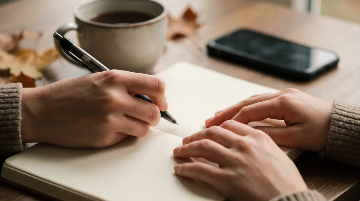 Close-up of young hands writing in a journal with a pen beside a cup of tea in soft daylight