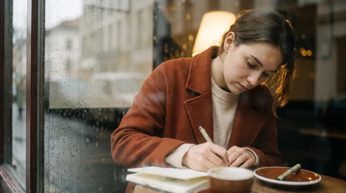 A woman writing in a notebook at a café window, coat still on, before anyone has read a word of her manuscript