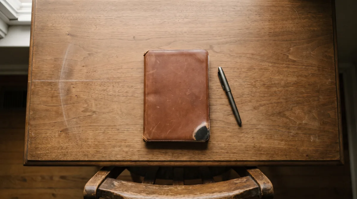 Overhead view of a closed notebook and a capped pen on a desk, chair edge just in frame
