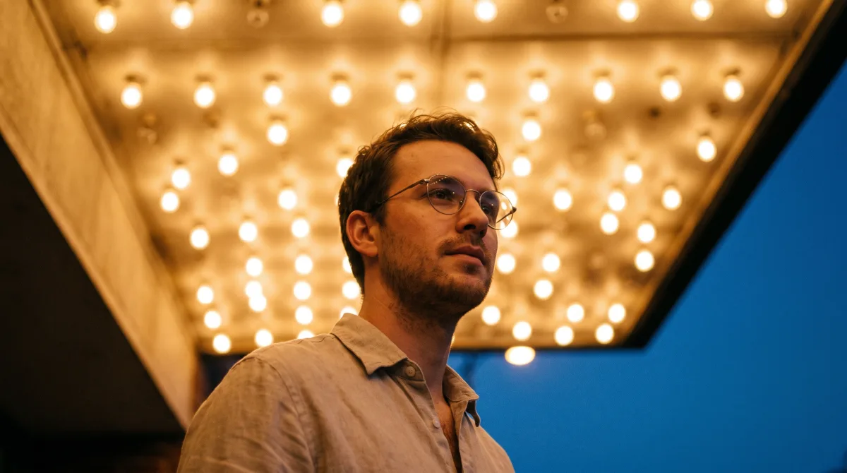 A man in his late twenties standing under a repertory cinema marquee at dusk, the marquee lights thrown fully out of focus behind him