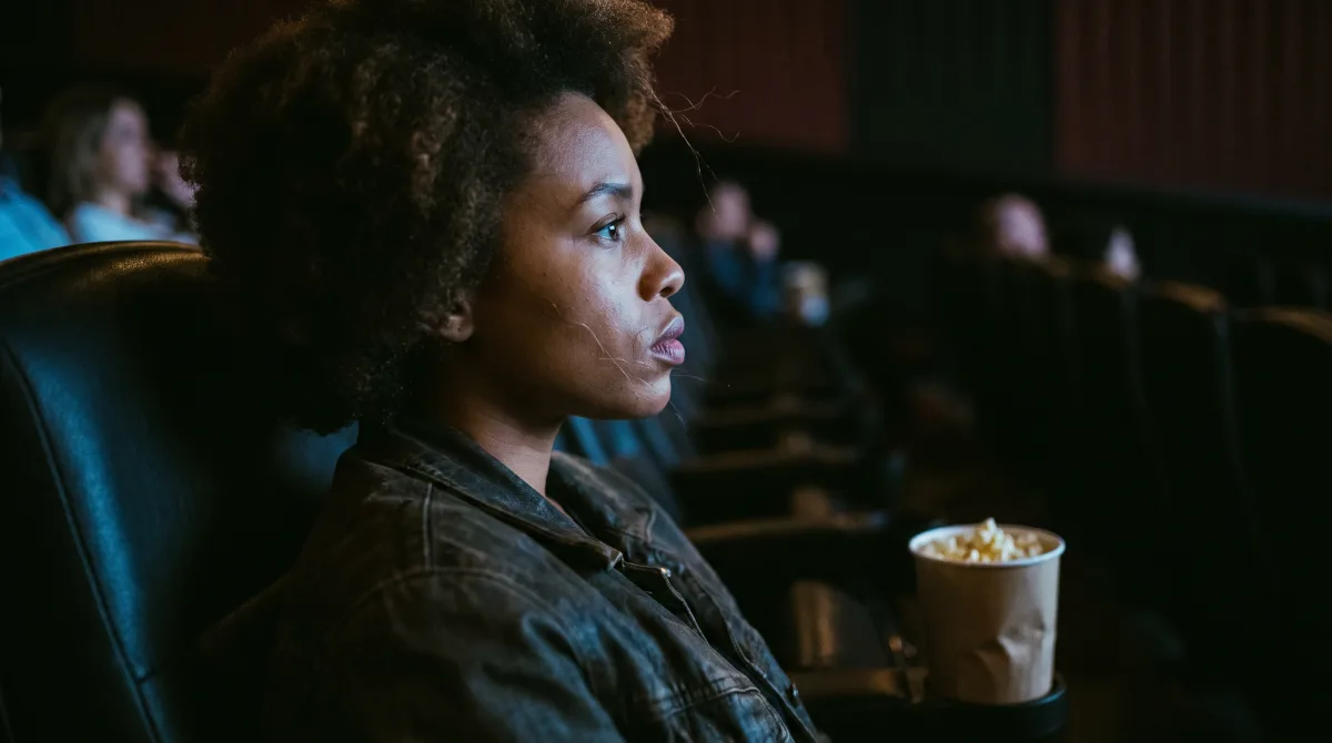 An 85mm profile portrait of a woman in a dark cinema seat, screen glow lighting her face, an untouched bag of popcorn on the armrest beside her