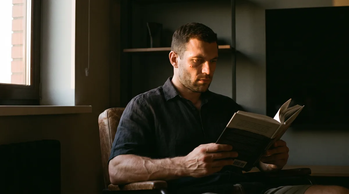 A man in his late 30s standing at a narrow window in a modern flat, listening intently with his eyes open