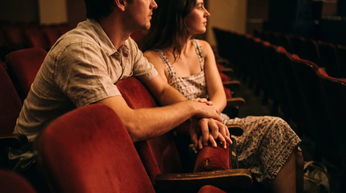 A man and a woman seated side by side in red velvet theatre seats, their hands touching on the armrest they share