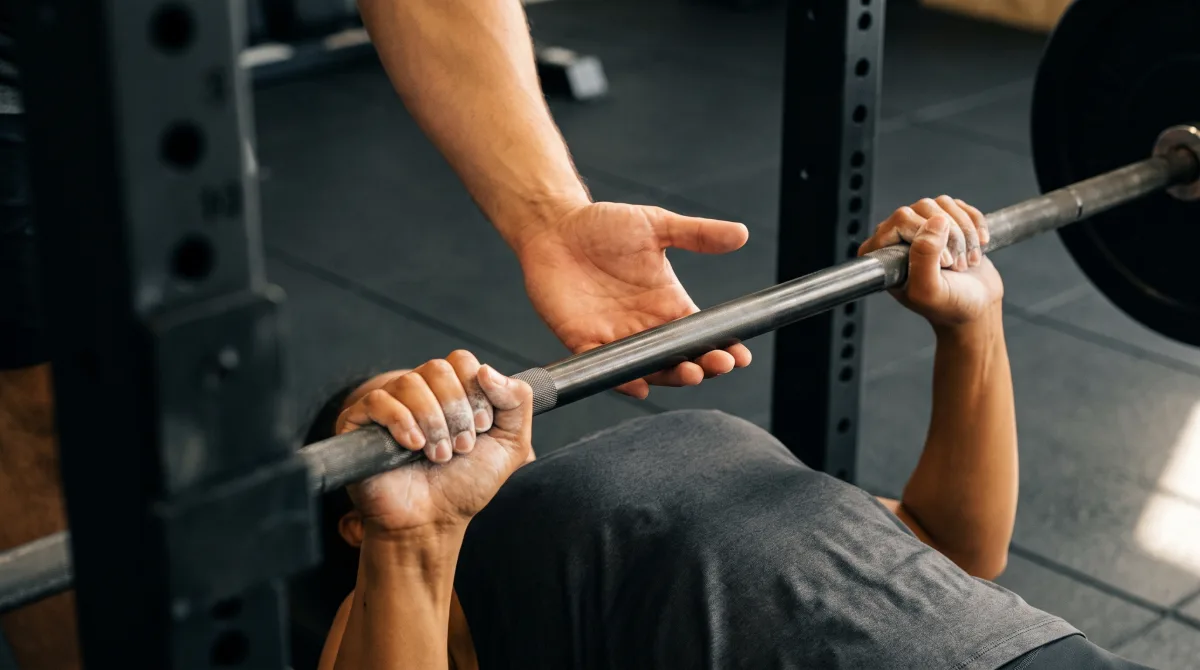 Close-up of one person's hand steadying a loaded barbell above another lifter's chest during a bench press