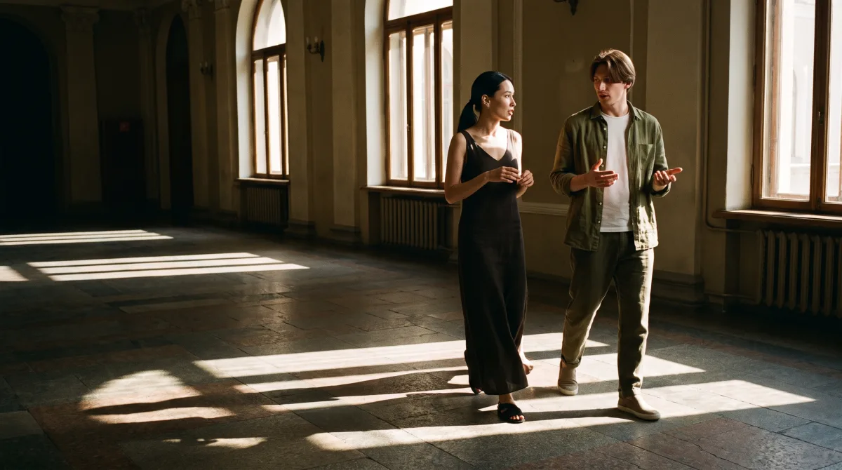 A couple mid-sentence stepping through a shaft of hard afternoon light in a theater foyer with tall glass windows