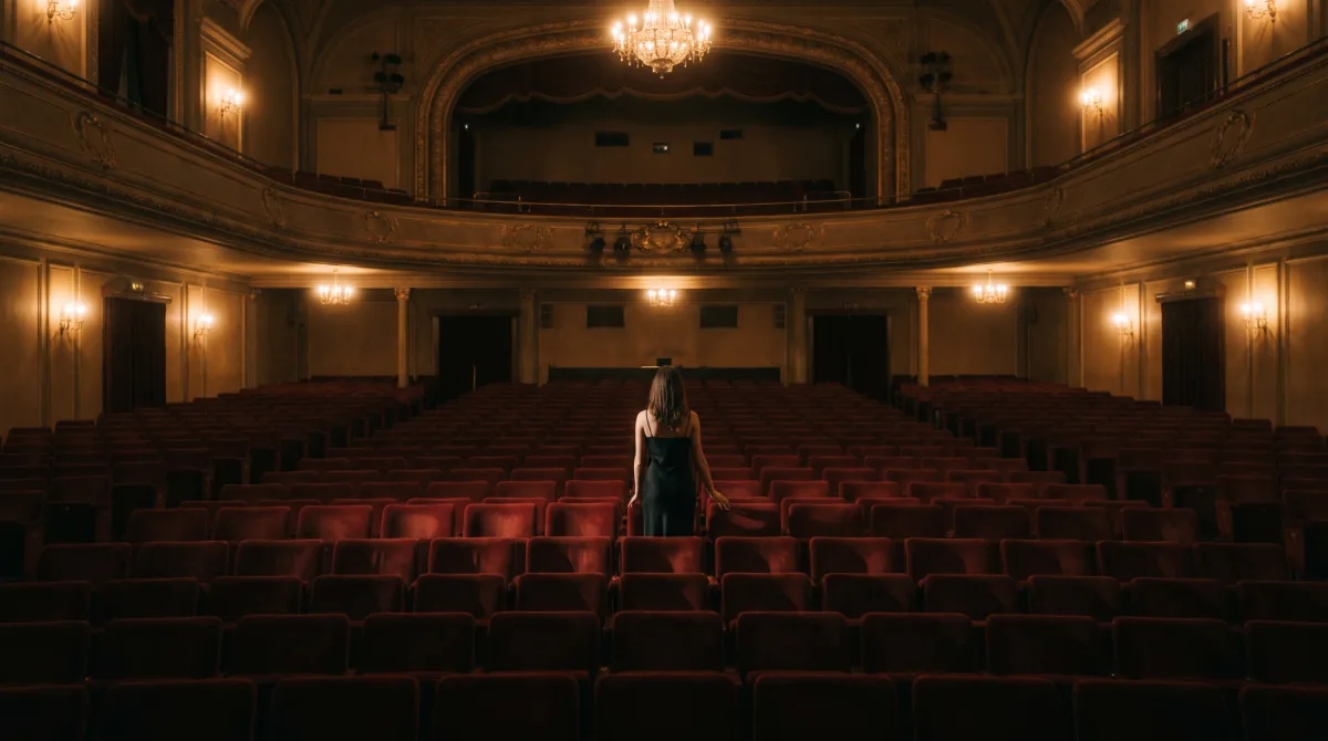 A person seen from behind taking a seat alone in an empty row of red velvet theater seats before the house lights go down