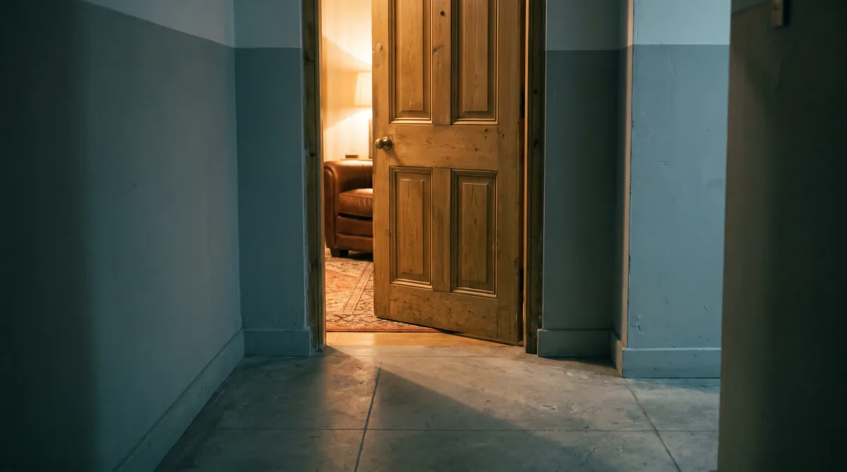 A door left ajar into a warm-lit room, seen from a cool, dim hallway — nobody in the frame