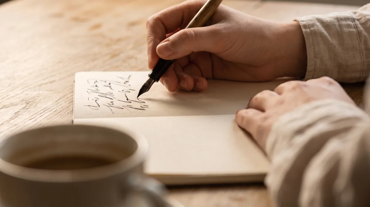 Close-up of a hand mid-word with a pen crossing a page on an oak table, the rim of a coffee cup at the edge of the frame