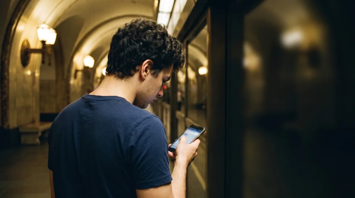 A young man on an evening metro platform reading his phone, his reflection faint in the dark tunnel-side window glass