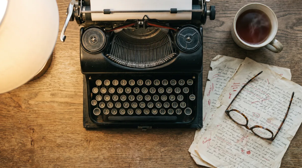 A vintage typewriter beside manuscript pages marked up in red editorial pencil, with reading glasses and tea — the close reading a literary editor gives a submission