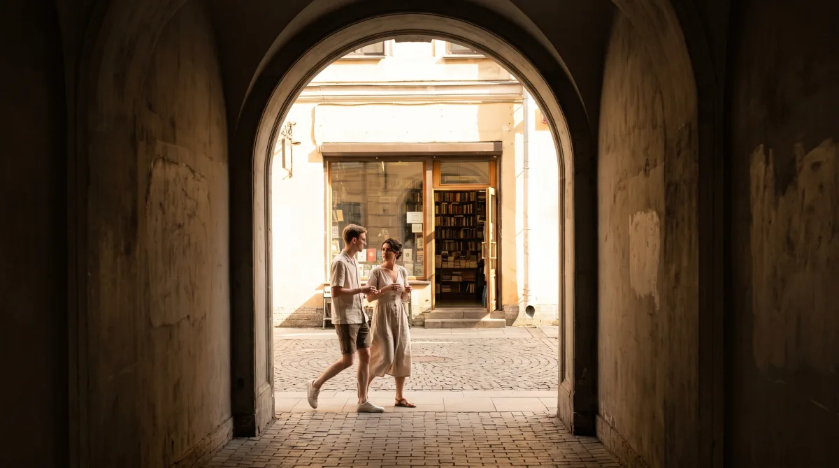 Two people mid-conversation walking from a covered arcade into a bookshop's warm interior light