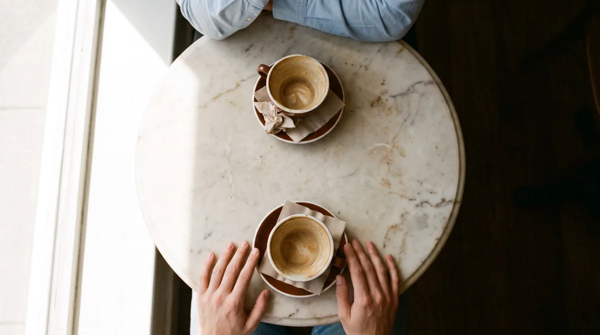 Two coffee cups on a marble café table with only one pair of hands in frame, a second person's cuff just cropped out of the shot