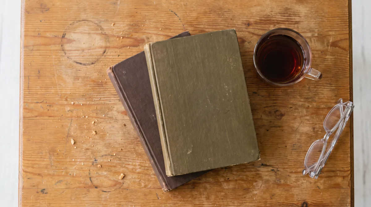 Two closed books stacked beside a half-full cup of tea on a worn wooden table, morning light crossing the grain